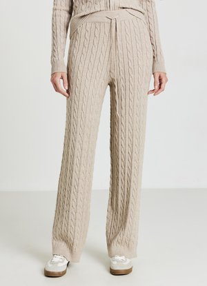 Beige weit geschnittene Strickhose mit Zopfmuster, ausgestattet mit einem enganliegenden Bund und einer Bindung. Kombiniert mit weißen und braunen Turnschuhen.