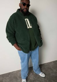Grön fleece zip-up jacka över en mörkgrön skjorta, ihop med ljusblå jeans och vita sneakers. Solglasögon fulländar outfiten.
