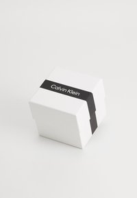 Scatola quadrata bianca con coperchio a cerniera, dotata di una fascia nera con il testo "Calvin Klein". Finitura liscia, senza ulteriori motivi o texture.