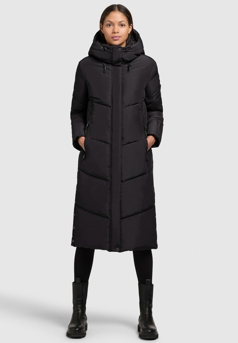 Długi czarny puchowy płaszcz z kapturem, z pionowymi przeszyciami, kieszeniami bocznymi i zapięciem na napy u dołu. Wykonany z pikowanego, ocieplanego materiału.
