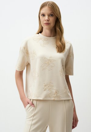 Beige T-shirt met korte mouwen en een reliëf bloemenpatroon, met ronde hals, gemaakt van zachte stof. Gecombineerd met lichtbeige broeken.