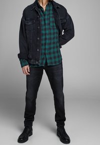 Chaqueta de mezclilla negra sobre una camisa de cuadros verde y negro, combinada con jeans oscuros y botas negras. Diseño simple en capas con detalles de botones.