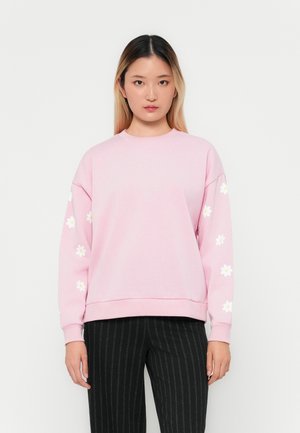 Jonge vrouw in een lichtroze sweatshirt met witte madeliefjesprints op de mouwen en zwarte streepjaspantalon, staande voor een witte achtergrond.