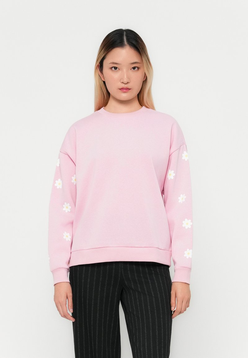 Ung kvinna som bär en ljusrosa sweatshirt med vita prästkragartryck på ärmarna och svarta ränderbyxor, står mot en vit bakgrund.