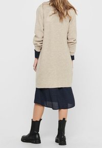 Beige pletený cardigan s dlouhými rukávy, spárovaný s modrými šaty s puntíky. Černé kotníkové boty dotvářejí outfit.