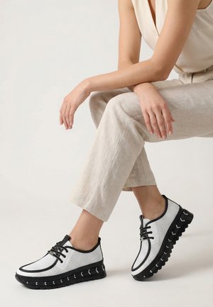 Femme portant un pantalon beige et un haut sans manches, assise, montrant des baskets blanches et noires à lacets avec des semelles épaisses.