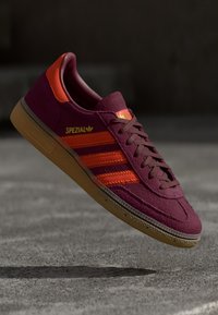 Maroon Adidas Spezial-sko med oransje striper og gummisåle, suspendert over grå betongoverflate i dempet belysning.