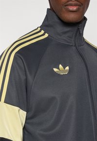 Tmavosivá športová bunda s žltými pruhmi, vysokým golierom, plným zipsom a vyšívaným zlatým logom na ľavej strane hrudníka.