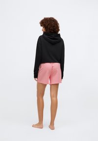 Zwarte cropped hoodie met capuchon, gecombineerd met roze shorts met een geometrisch patroon. Model staat blootsvoets, met de rug naar de camera.