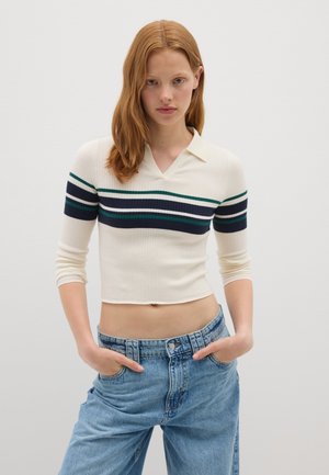 Giovane donna con i capelli rossi che indossa un maglione corto bianco con strisce navy e verdi e jeans chiari a vita alta, con le mani nelle tasche.