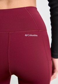 Burgundy leggings i stretchtyg, hög midja med en ficka, med en vit Columbia-logotyp nära midjebandet och en slät yta.