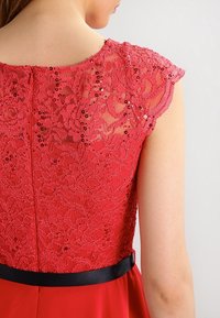 Robe rouge avec un corsage en dentelle à motifs floraux et paillettes. Le dos comprend une fermeture éclair dissimulée et une ceinture en satin noir à la taille.