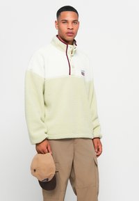 Light green fleece pullover with a high collar, maroon accents, and a small logo patch. Model holds a beige cap with a darker brim.