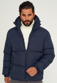 Marineblaue Steppjacke mit Kapuze, ausgestattet mit Reißverschluss, gerippten Bündchen und horizontalen Steppmuster-Details. Getragen über einem cremefarbenen Strickpullover.