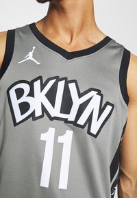 Maglia da basket grigia con rifiniture nere, caratterizzata da un grande testo bianco "BKLYN" e dal numero "11" sul davanti; realizzata in materiale in rete traspirante.