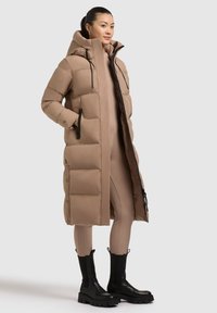 Manteau doudoune beige long avec un col haut, fermeture éclair et deux poches latérales. Présente un design matelassé et des accents en métal noir.
