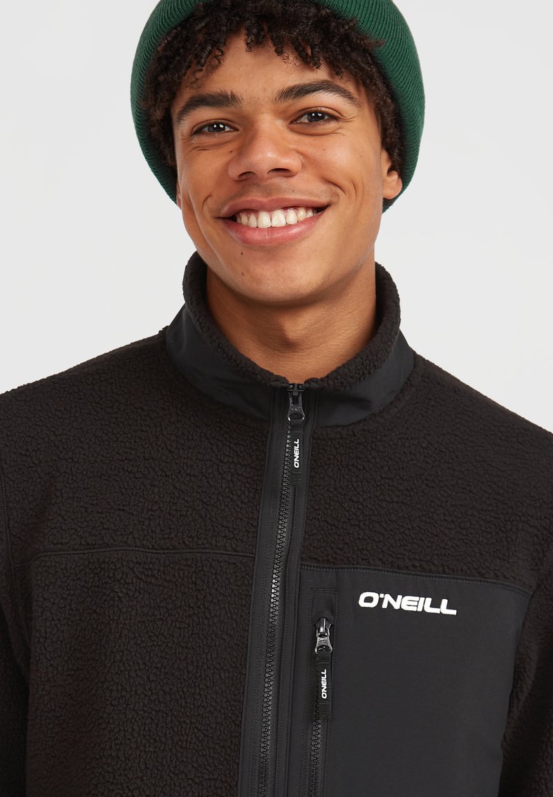 Giovane sorridente che indossa un berretto verde e una giacca in pile nera O'Neill con tasca a zip sul petto, su uno sfondo semplice.