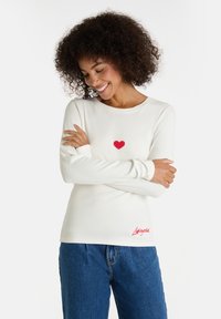 Frau mit lockigem Haar, die ein weißes Langarmshirt mit einem roten Herz und rotem Text trägt, die Arme verschränkt, lächelt und nach unten schaut.