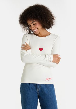 Frau mit lockigem Haar, die ein weißes Langarmshirt mit einem roten Herz und rotem Text trägt, die Arme verschränkt, lächelt und nach unten schaut.