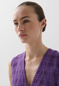 Rochie cu decolteu în V, dintr-un material texturat de culoare mov cu modele în carouri. Prezintă o croială lejeră și pliuri subtile în jurul decolteului și umerilor.