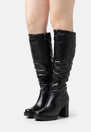 Bottes en cuir noires montantes jusqu'aux genoux avec talons carrés et fermetures éclair sur le côté, portées sur des jambes nues sur un fond blanc uni.