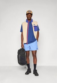 Fleecevest med beige og blå detaljer, blå skjorte, lyseblå shorts, svarte sko og en svart ryggsekk. Kjennetegn inkluderer fargekontrast og lommer.