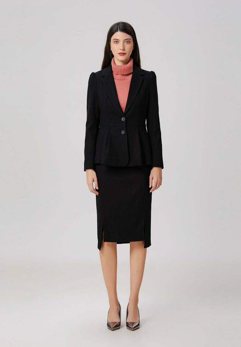 Femme se tenant debout, portant un blazer noir sur un col roulé rose et une jupe noire arrivant aux genoux avec des fentes, assortie à des talons métalliques pointus.