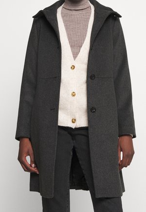 Mørkegrå knælange frakke over beige cardigan med knapper og lysebrun ribstrikket rullekrave, båret med sorte bukser.