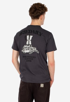 Mann trägt ein schwarzes T-Shirt mit einer Schildkröte, die eine Mokkakanne trägt, und den Texten "IRIEDAILY" und "Slowpresso Coffee Club" auf dem Rücken.