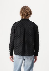 Black fabric shirt with raised black dot texture, long sleeves, loose fit, and buttoned back yoke detail, paired with light blue jeans.