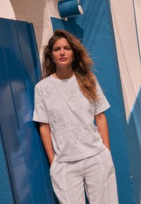 Biele vyšívané tričko s kvetinovými vzormi, doplnené voľnými bielymi ľanovými nohavicami, na pozadí modro-bielej steny.