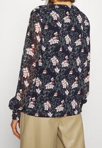 Blumenbluse aus transparentem, leichten Stoff. Dunkelblauer Hintergrund mit pinken und weißen Blumen sowie grünen Akzenten. Gummizüge an den Ärmeln. Rundhalsausschnitt.