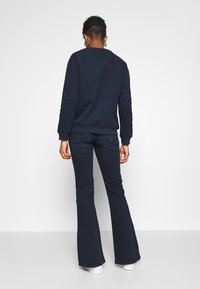 Person med kort lockigt hår som bär en marinblå sweatshirt, mörka utsvängda jeans och vita sneakers, stående vänd mot en vit vägg.