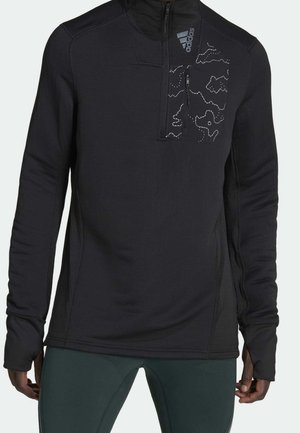 Schwarzer langärmliger Pullover mit Viertelreißverschluss, Adidas-Logo und gemusterter Brusttasche, getragen von einer Person mit grüner Hose.
