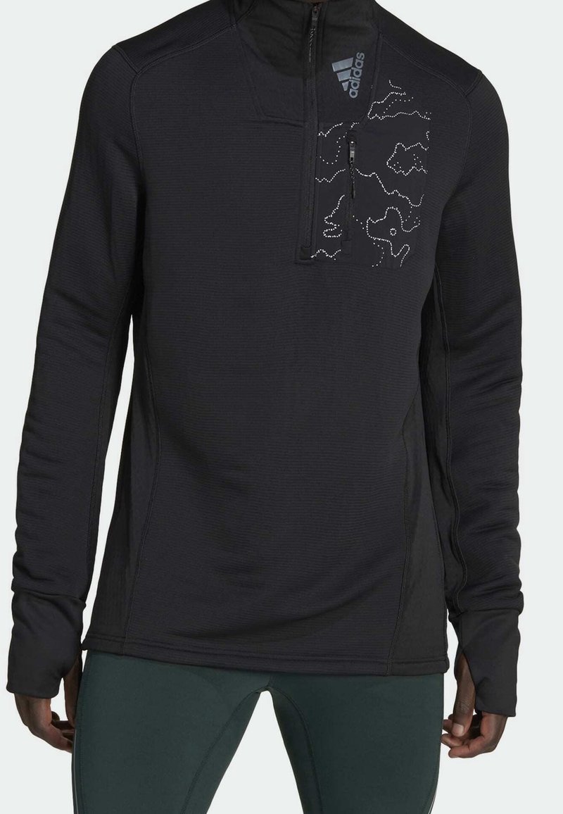 Pull noir à manches longues avec fermeture éclair quart de zip, logo Adidas et poche poitrine à motifs, porté par une personne avec un pantalon vert.