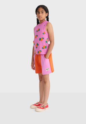 Meisje met gevlochten haar, gekleed in een roze mouwloos topje met oranje ananassen, oranje en roze korte broek, en rode schoenen met bloemenmotieven.