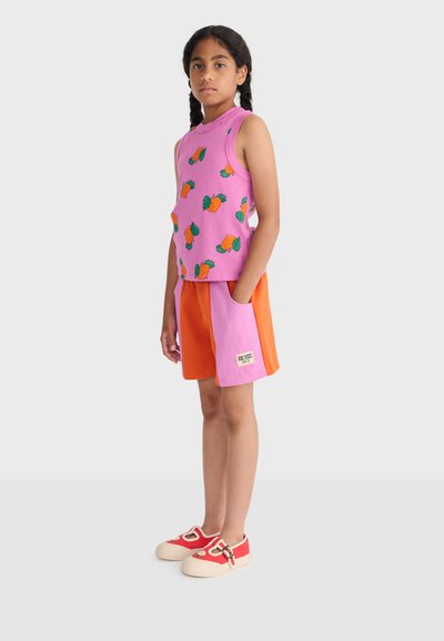 Mädchen mit geflochtenem Haar, das ein pinkfarbenes ärmelloses Oberteil mit orangefarbenen Ananas, orange-rosa Shorts und rote Schuhe mit Blumendetails trägt.
