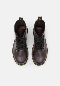 Un par de botas lace-up de un brillante burdeos oscuro con costuras amarillas alrededor de suelas negras, vistas desde arriba sobre un fondo blanco.