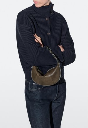 Person wearing a navy ribbed sweater and dark jeans, holding a brown leather crescent-shaped handbag with a chain strap across their forearm.