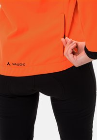 Orange jacka med en dragkedja på sidan, tillverkad av ett smidigt och lätt material. Svarta detaljer vid ärmsluten och den nedre fållen. Logotyp synlig.