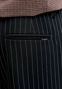 Gros plan sur la poche d'un pantalon à rayures noires avec une petite étiquette indiquant "JACK & JONES" et une partie d'une chemise à carreaux marron au-dessus.