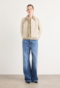 Beige strukturierte Jacke mit Kragen und Knöpfen, kombiniert mit hellblauen, weiten Jeans und schwarzen spitzen Schuhen.
