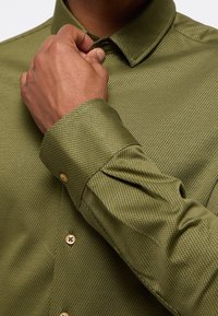 Camicia verde a fantasia con colletto button-down, maniche lunghe e bottoni dorati a contrasto lungo la parte frontale e sui polsi.