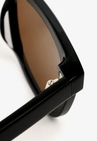 Gafas de sol negras con un marco rectangular, lentes marrones y un grabado visible de "SUPERDRY" en la varilla, con acentos metálicos.