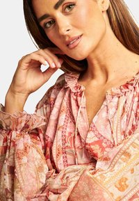 Bluse mit pinkem Blumenmuster, Rüschensaum am Ausschnitt und an den Ärmelabschlüssen, mit unterschiedlichen Texturen und Farben in einem fließenden Design.