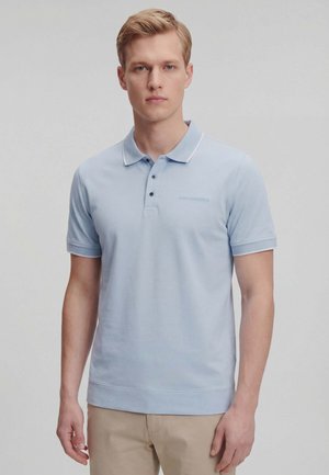 Joven con cabello rubio corto, vestido con una camisa polo azul claro y pantalones beige, de pie frente a un fondo gris liso.