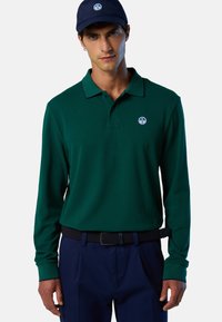 Polo de manga larga en algodón verde oscuro con cuello clásico y parche con logo. Combinado con pantalones azul marino y una gorra a juego.