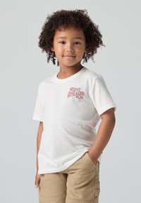 Balta medvilninė marškinėliai trumpomis rankovėmis su rožiniu Levi Strauss & Co. logotipu kairėje krūtinės pusėje, derinami su smėlinėmis šortais.