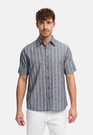 Mand iført en grå kortærmet skjorte med hvide lodrette striber og hvide bukser, stående foran en ensfarvet baggrund.