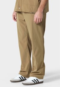 Pantalones rectos, ligeros y de color beige, con una textura suave, complementados por un bolsillo y usados con zapatillas blancas con rayas negras.